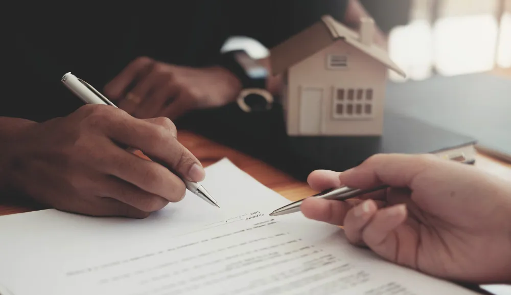 Mãos assinando contrato com miniatura de casa, pensando em como evitar que filhos vendam imóveis.